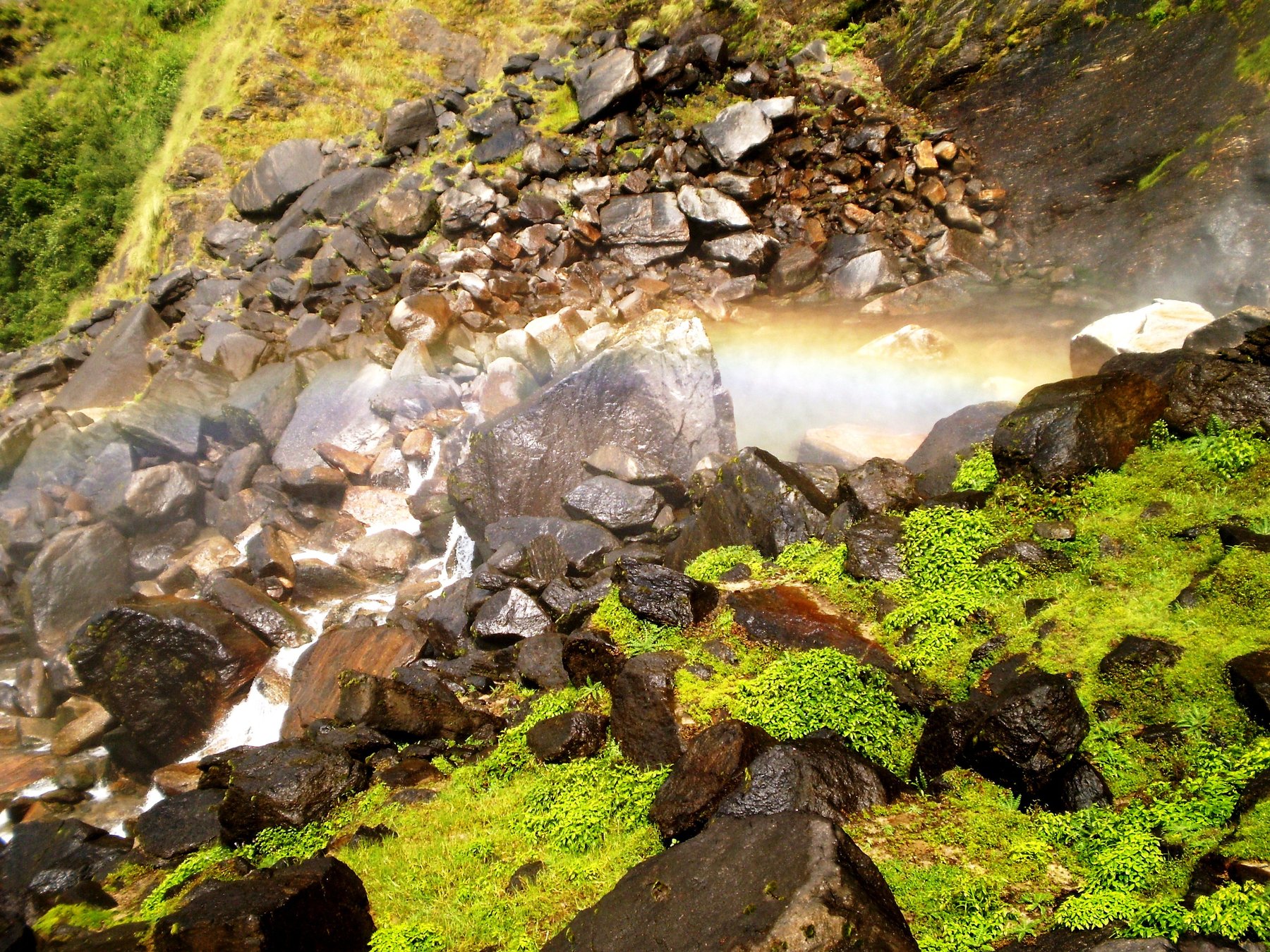 A rainbow forming in the mist over the wet boulders at the base of Birthi Falls