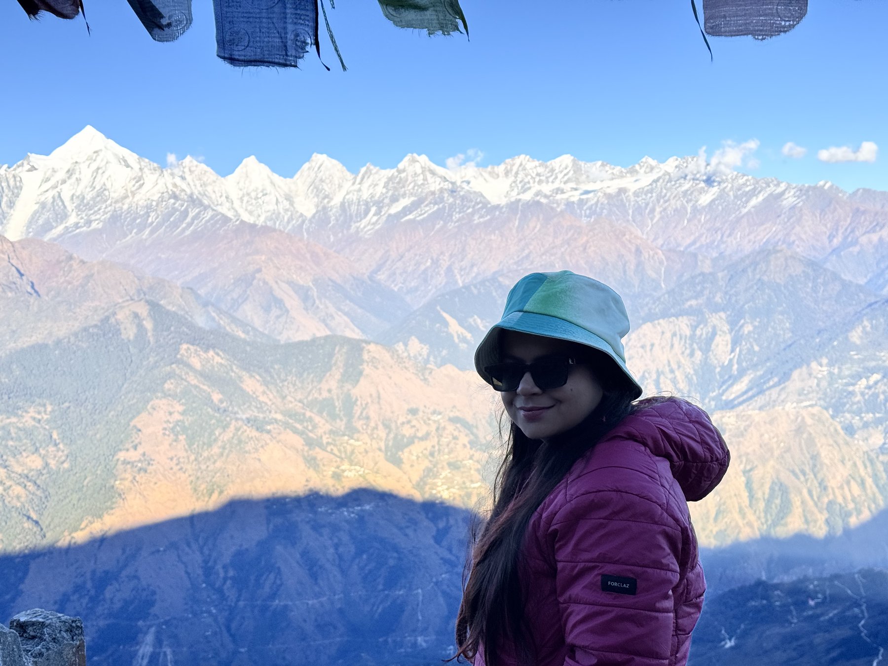 A trekker in a bucket hat smiling beside prayer flags, with the golden-lit Panchachuli range behind