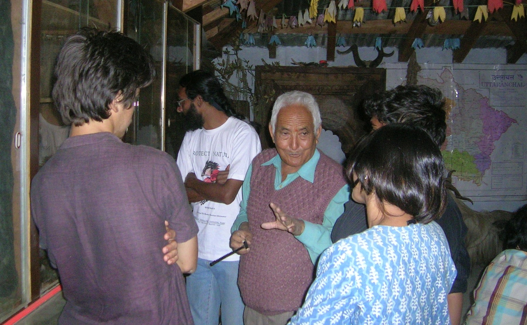 Dr. S.S. Pangtey explaining exhibits to visitors inside his tribal heritage museum, prayer flags and a wall map of Uttarakhand behind him